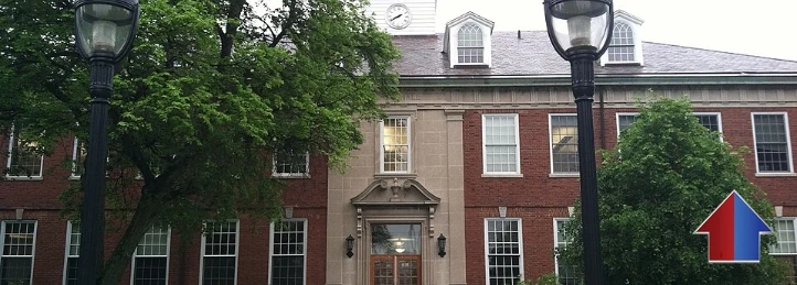 Historic red brick academic building with tall windows, trees, and lampposts on a college campus.