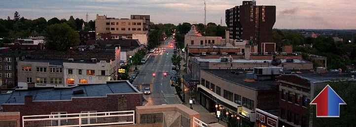 Downtown city street at dusk with storefronts, office buildings, and traffic lights viewed from above.